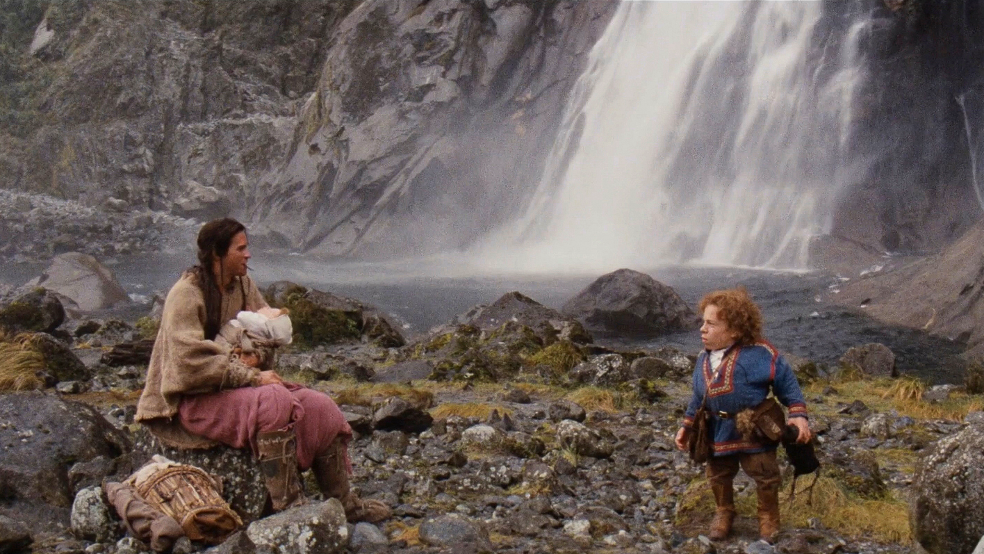 On a rocky shore beneath a waterfall, a man with light skin sits and holds a baby by another man with light skin who has dwarfism and curly red hair.