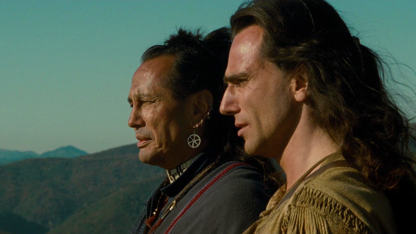 A close still of two men in profile looking out over a landscape. An older man with medium skin and long dark hair and a younger man with light skin.