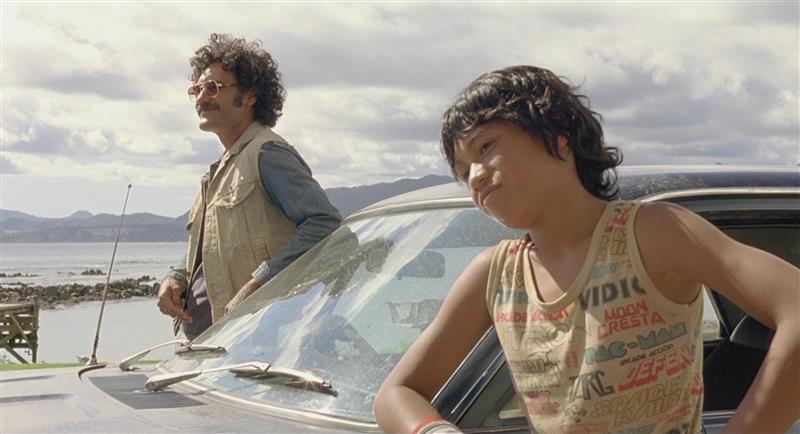 A boy in a tank top leans against a car. A man stands on the opposite side wearing sunglasses and a vest. Both have dark curly hair and medium skin tone.