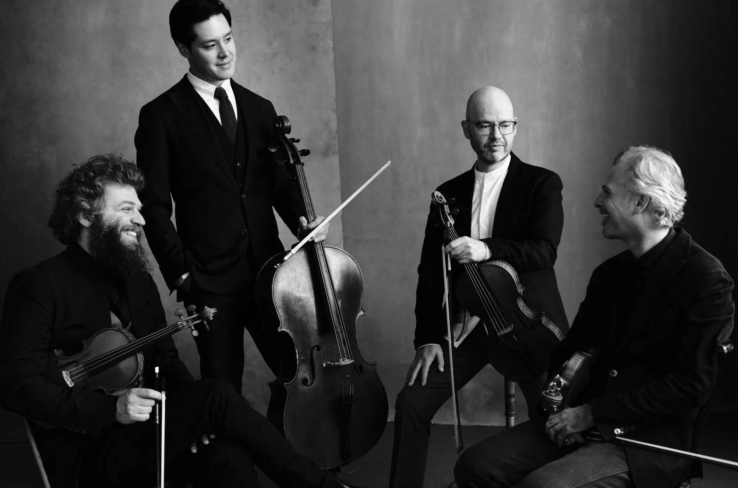 Four men in formal attire, holding string instruments (two violins, a viola, and a cello), sit and stand together smiling and conversing in a studio with a plain backdrop. The image is in black and white.