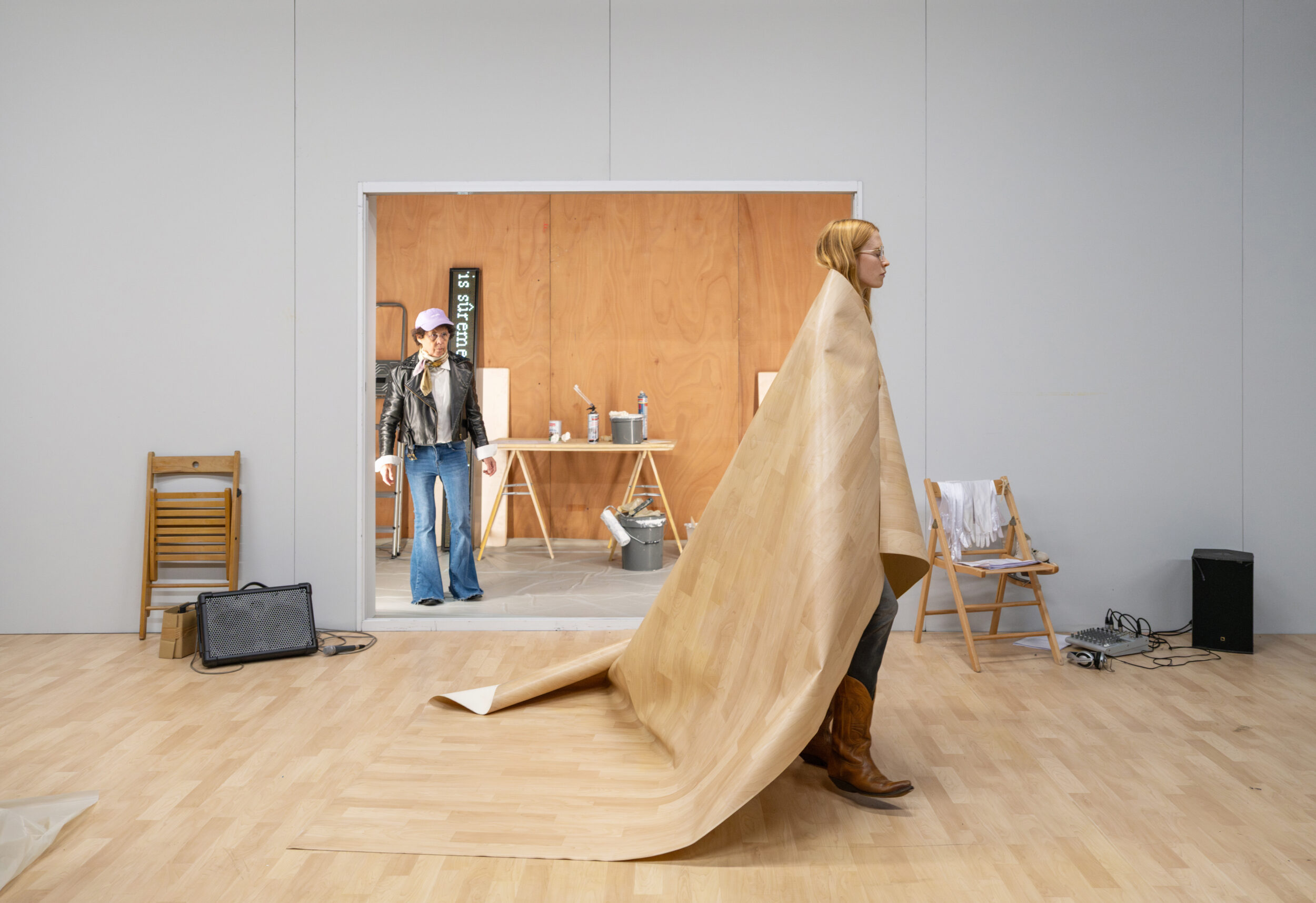 A person in boots walks wrapped in a rolled-up sheet of flooring, while another person stands in a small, modern room in the background. Folding chairs and scattered objects are visible on the floor.
