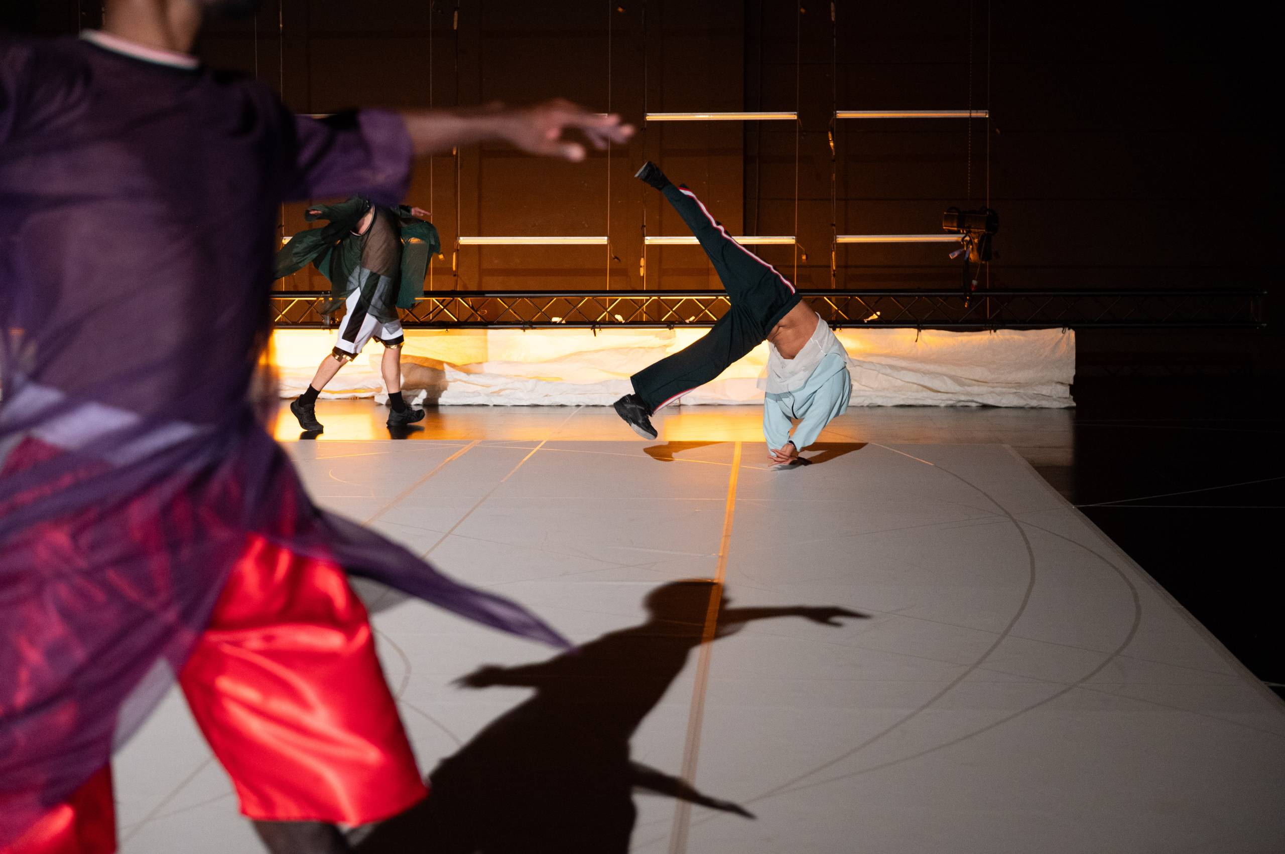 A dancer performs a one-handed handstand on a lit stage while another dancer in red shorts and a sheer purple top moves in the foreground, both casting shadows on the floor.
