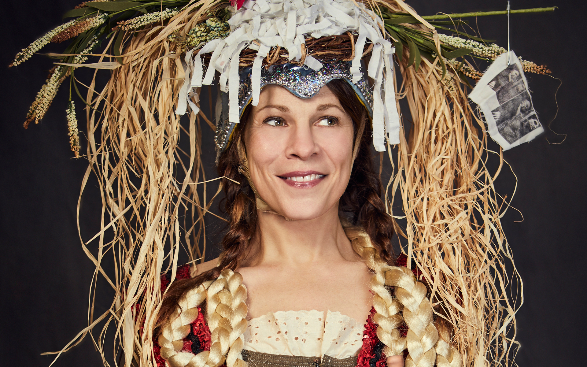 A woman with long blonde braids wears a decorative headdress made of straw, paper, flowers, and photographs, smiling slightly and looking off to the side against a dark background.
